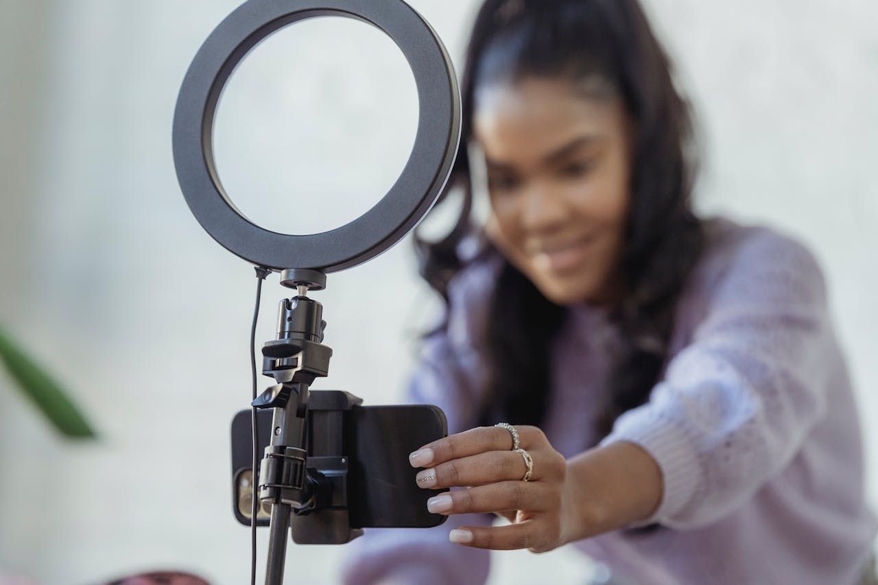 journey-02 Cheerful young African American female blogger in stylish sweater smiling while setting up camera of smartphone attached to tripod with ring light before recording vlog